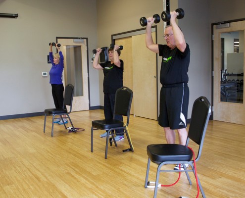 people performing shoulder press with dumbbells in gym room