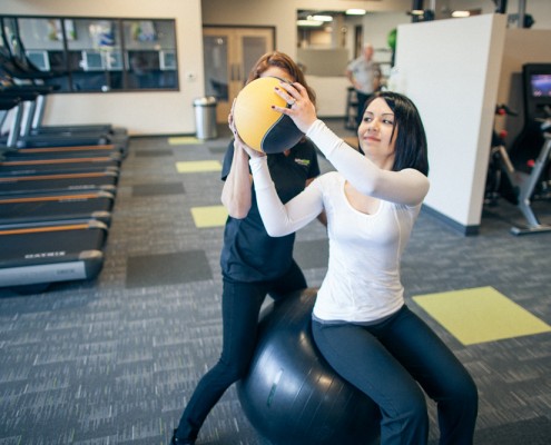 female personal trainer assists female member seated on exercise ball lifting weighted ball
